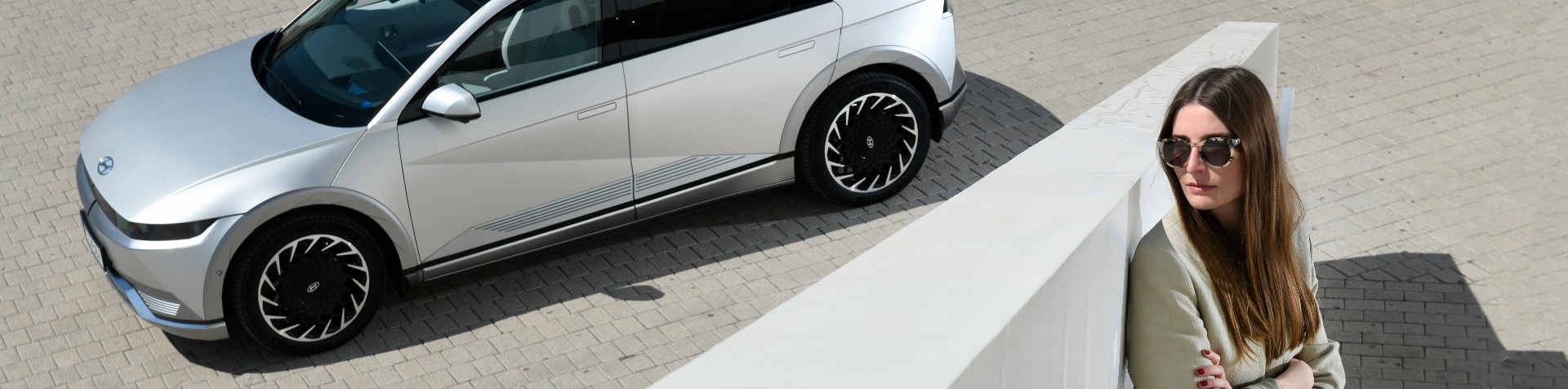 Britta Reineke sitting behind a wall in front of a gray IONIQ 5. She has long brunette hair and is wearing large tortoise shell sunglasses and a beige blazer. 