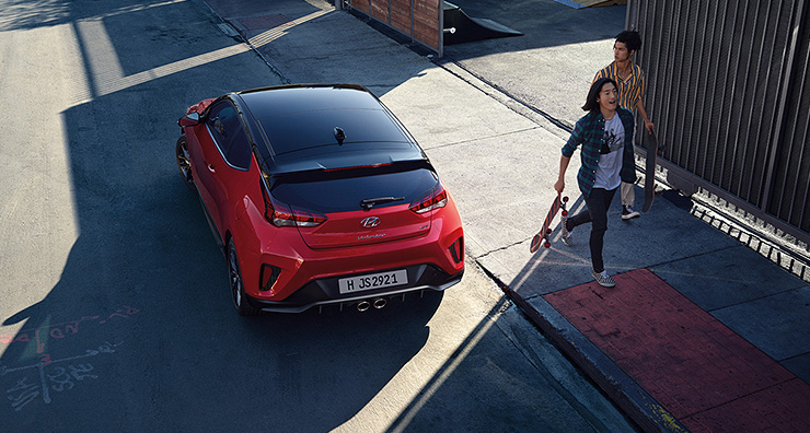 Red Veloster parked on the road beside persons.