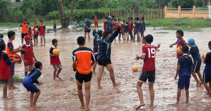 Lots of kids wearing red and blue jerseys are playing football in a rainy day, with yellow color balls.