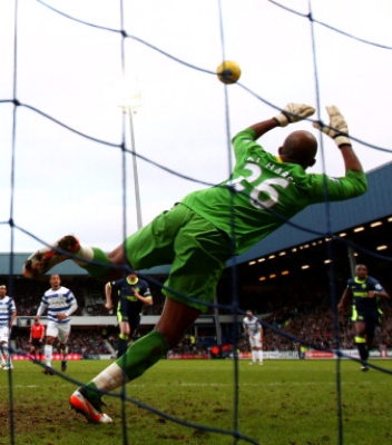 Action picture taken from behind the net of Team Century male footballer Ali Al-Habsi in green game jersey with the number 26 on the back jumping almost horizontally to stop a yellow ball.